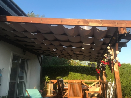 Beige premium waterproof Roman blind installed under a wooden pergola on a sunny backyard terrace holding outdoor furniture.