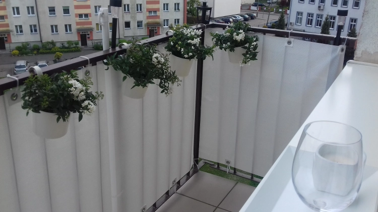 White balcony privacy screen mounted on a railing with hanging white flower pots and a city view.