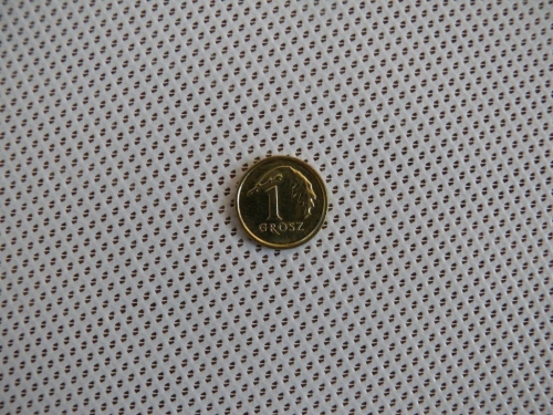Single 1 Grosz Polish coin placed on a white perforated Roman sun shade fabric with small triangular holes