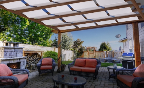 Outdoor pergola with a white sunshade canopy over a stone patio with wicker furniture and a fireplace