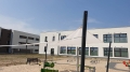 Háromszögletű bézs napvitorla 3,2x3,2x4,5 m homokozó felett egy játszótéren egy modern épület mellett