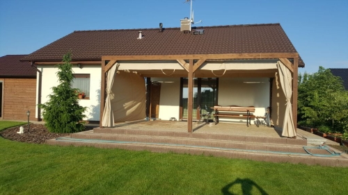 Beige waterproof garden curtains hung on a custom wooden pergola attached to a modern house with a patio.
