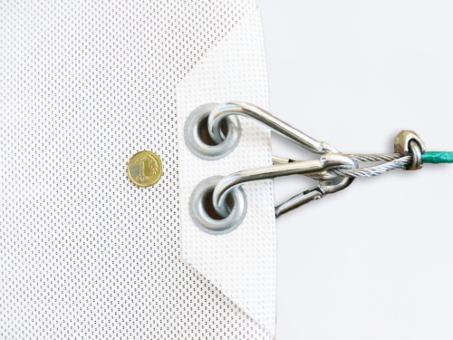Close-up of a white sun sail corner with metal grommets, carabiners, and a steel cable next to a small coin for scale