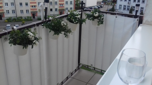 White woven privacy screen installed on a balcony railing with hanging flower pots and a city view.